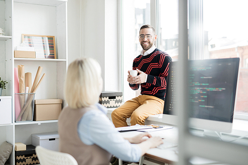 Cheerful excited handsome office man in stripped sweater sitting on window sill and drinking coffee while talking to coder typing on computer keyboard
