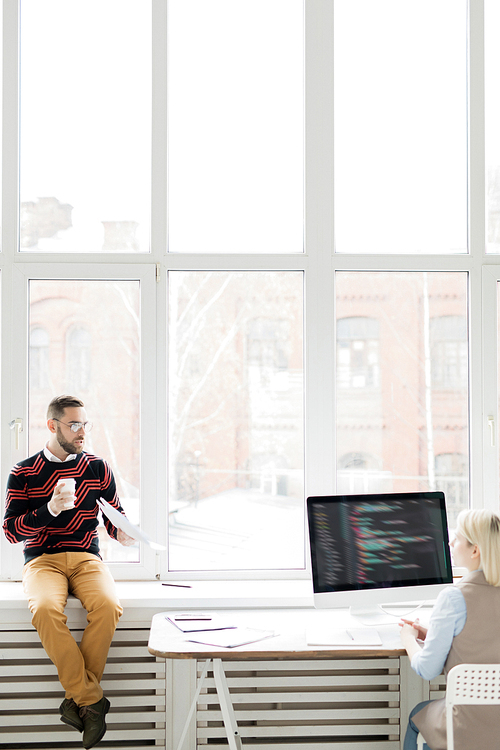 Serious displeased handsome bearded manager in stylish sweater sitting on window-sill and gesturing hand with papers while reporting employee working with computer language
