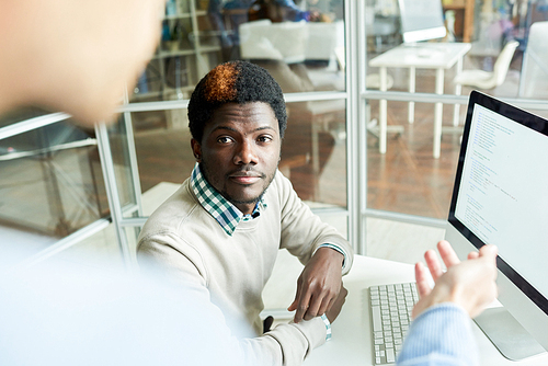 Portrait of young African office worker sitting at desk in office and listening to colleague standing beside him in discussion of business project