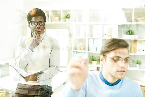 Portrait of two IT specialists writing formulas on glass wall in modern office, focus on young African-American man wearing glasses, copy space