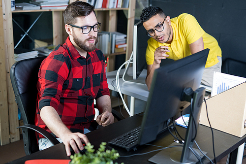 Portrait of two modern young men wearing glasses using computer and discussing projects in IT office of creative developers team, copy space