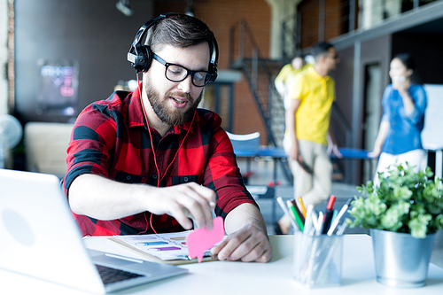 Portrait of modern bearded man wearing headphones and glasses  sitting at desk in modern office of IT developers company, copy space