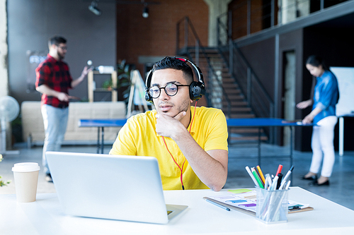 Portrait of  creative young Middle-Eastern man wearing glasses and bright yellow shirt using laptop while working in open space office of IT developers team, copy space