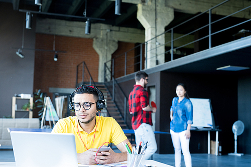 Portrait of  creative young Middle-Eastern man wearing headphones and bright yellow shirt using laptop and listening to music while working in open space office of IT developers team, copy space