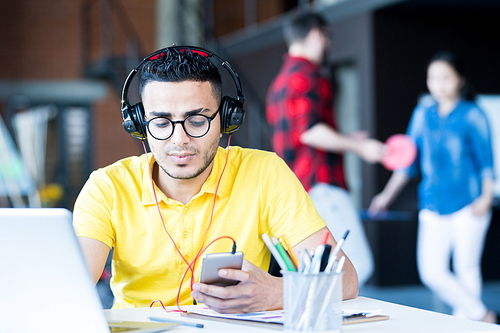 Portrait of  creative young Middle-Eastern man wearing headphones and bright yellow shirt using laptop and smartphone while working in open space office of IT developers team, copy space