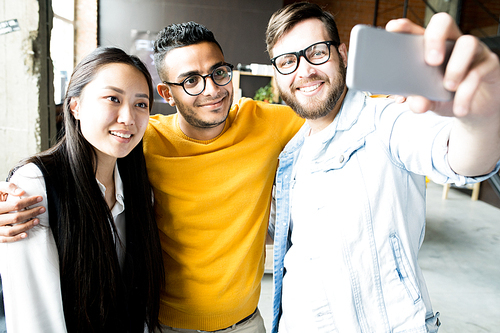 Multi-ethnic group of three creative young people smiling happily while taking selfie via smartphone standing in modern office