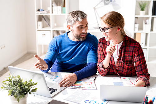 Cheerful optimistic young colleagues in casual clothing sitting at table and discussing marketing analysis in office: bearded man pointing at screen while explaining strategy to woman in earphones
