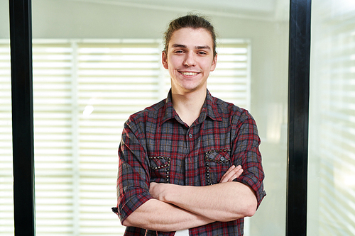 Waist-up portrait of young Caucasian man in casual shirt standing in office with his hands crossed and smiling at camera happily