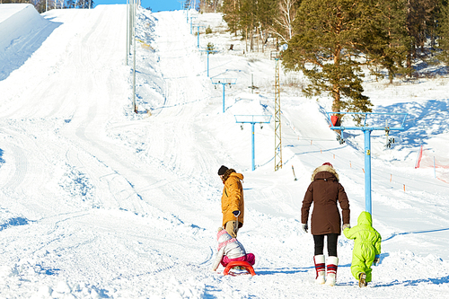 Rear view of parents with little kids going towards ski slope on beautiful winter day, father dragging sled with his daughter