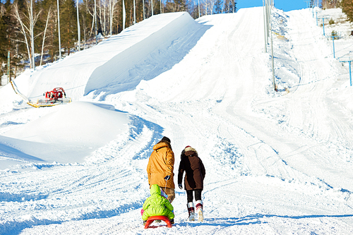 Rear view of couple walking towards snow slope and pulling sled with their little son on sunny winter afternoon