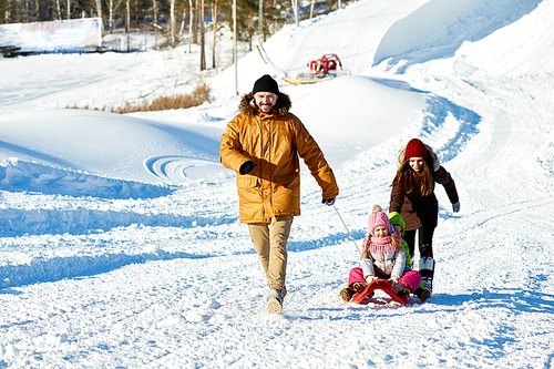 Caucasian man dragging sled with little children along snowy road on frosty sunny day, his wife walking behind them