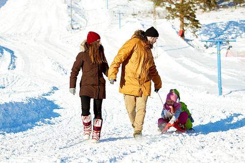 Caucasian couple holding hands while walking and dragging sled with their little kids on beautiful winter afternoon out of town