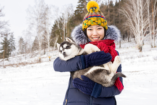 Waist up portrait of smiling Asian woman holding cute Husky puppy  while posing against winter landscape, copy space