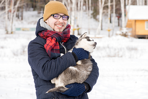 Waist up portrait of modern young man smiling at camera while holding cute Husky puppy outdoors in winter, copy space