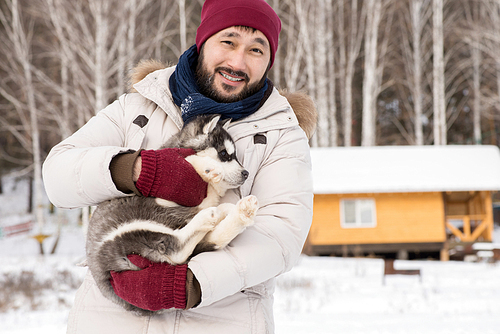 Waist up portrait of modern Asian man smiling at camera while holding cute Husky puppy outdoors in winter, copy space