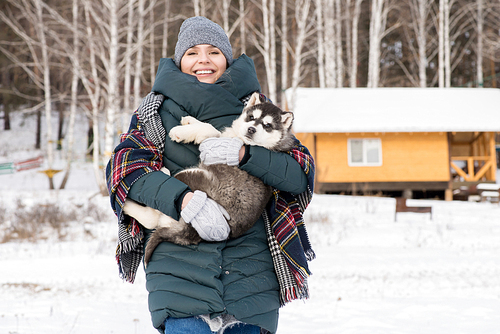 Waist up portrait of modern young woman smiling at camera while holding cute Husky puppy outdoors in winter, copy space