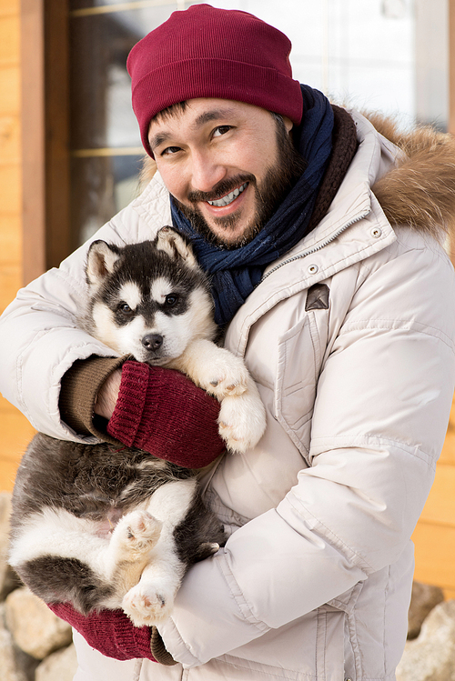 Portrait of cheerful Asian man holding puppy  and smiling happily while posing outdoors on winter day