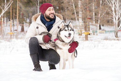 Full length portrait of modern Asian man posing with gorgeous Husky dog sitting down outdoors in winter landscape and smiling happily, copy space