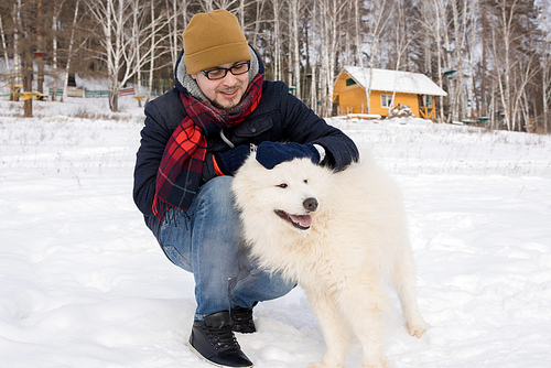 Full length portrait of modern young man playing with gorgeous Samoyed dog sitting down outdoors in winter landscape and smiling happily, copy space