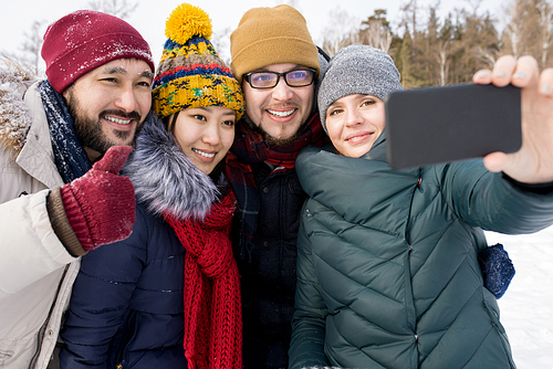 Waist up portrait of four happy young people taking selfie photo and showing thumbs up while having fun outdoors in winter