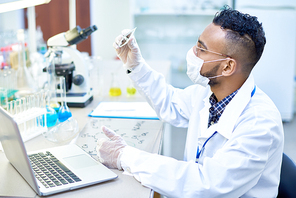 Side view portrait of young Middle-Eastern scientist working with test tubes and laptop studying bacteria in modern medical laboratory