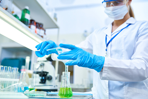 Portrait of female scientist wearing protective gloves pouring colored liquids to  test tubes while studying chemicals in medical laboratory, focus on hands