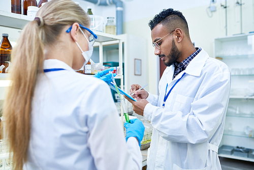 Side view portrait of young Middle-eastern scientist taking notes on clipboard while working on medical research in laboratory with his female colleague