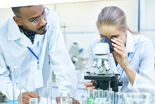 Portrait of young Middle-eastern scientist taking notes on clipboard while working on medical research in laboratory while his female colleague looking in microscope