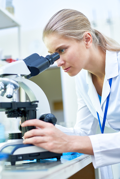 Side view portrait of young female scientist looking in microscope while working on medical research in science laboratory