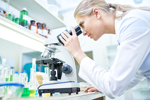 Side view portrait of young female scientist looking in microscope while working on medical research in modern laboratory