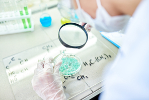 Close-up of unrecognizable scientist in protective gloves using magnifying glass while viewing small crystal joint with tweezers at table with glass board