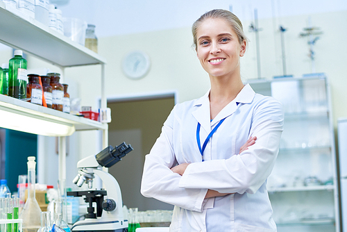 Portrait of young female scientist smiling cheerfully and  while posing in modern laboratory with arms crossed, copy space