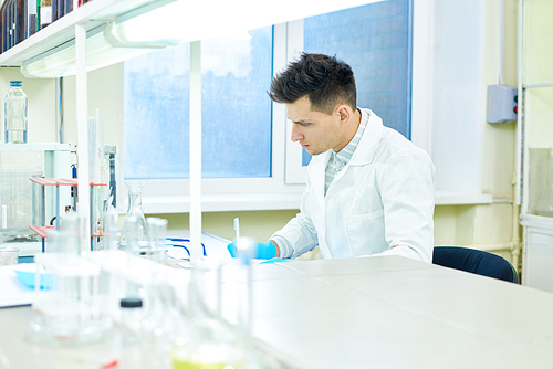 Profile view of handsome young researcher wearing rubber gloves and white coat sitting at desk and taking notes while carrying out experiment in modern lab