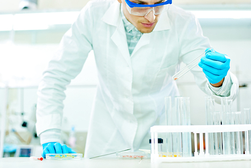 Handsome young researcher wearing safety goggles and white coat looking at test tube with concentration while carrying out experiment in modern lab