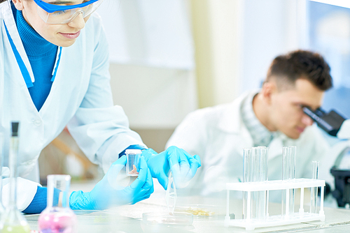 Pretty young scientist wearing safety goggles and white coat standing at lab bench and inspecting artificial meat sample while working on innovative scientific project with male colleague.