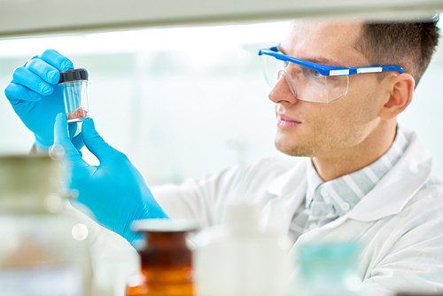 Concentrated young researcher wearing safety goggles and rubber gloves inspecting artificial meat sample while working on innovative scientific project