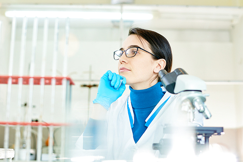 Waist-up portrait of pensive young scientist wearing eyeglasses and white coat looking away while preparing report about conducted experiment, interior of modern lab on background