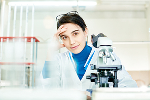 Waist-up portrait of attractive young scientist wearing white coat  with warm smile while sitting at desk in modern laboratory