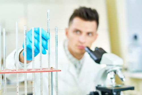 Young researcher wearing white coat and rubber gloves sitting in front of microscope and taking graduated pipette from holder, focus on foreground