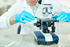 Close-up shot of unrecognizable male researcher wearing white coat and rubber gloves examining sample with microscope while sitting at laboratory desk