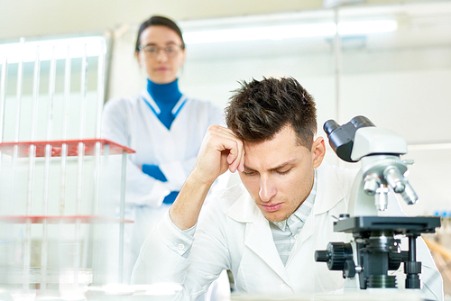 Concentrated young researcher wearing white coat leaning head on hand while sitting at desk, his pretty female colleague standing behind him, interior of modern lab on background