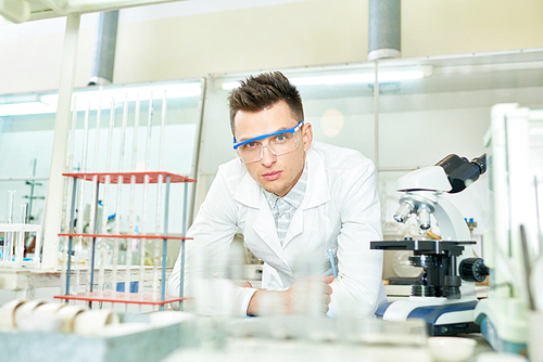 Handsome young medical technician wearing safety goggles and white coat leaning on laboratory bench and , portrait shot