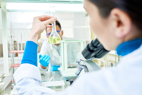 Over shoulder view of young dark-haired chemist holding flask with yellow solution in hand while sitting at desk, interior of modern lab on background