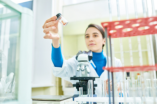 Confident young researcher wearing white coat sitting at desk and inspecting sample of in vitro meat, microscope and laboratory glassware on foreground