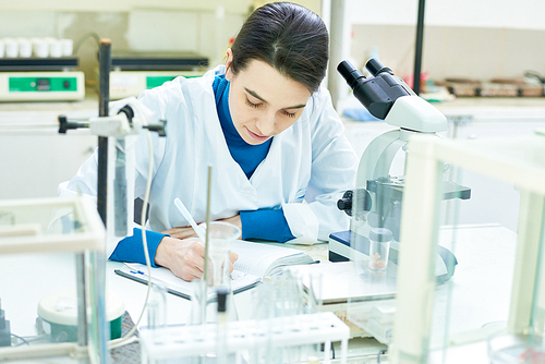 Beautiful dark-haired researcher wearing white coat sitting at desk and taking notes after successful completion of scientific experiment, interior of modern laboratory on background