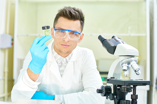 Waist-up portrait of confident young researcher wearing safety goggles and white coat sitting at desk of modern lab with GMO lettuce sample in hand and .