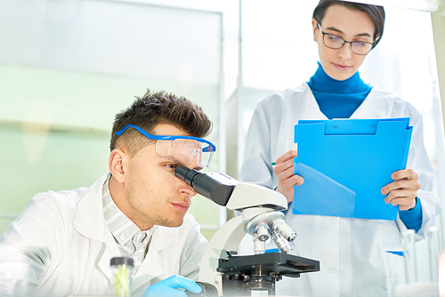 Handsome young scientist wearing safety goggles and white coat looking through microscope and his pretty colleague taking necessary notes while they carrying out experiment