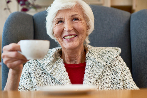 Head and shoulders portrait of cheerful elderly woman with charming smile looking away while enjoying delicious cappuccino at cozy small coffeehouse