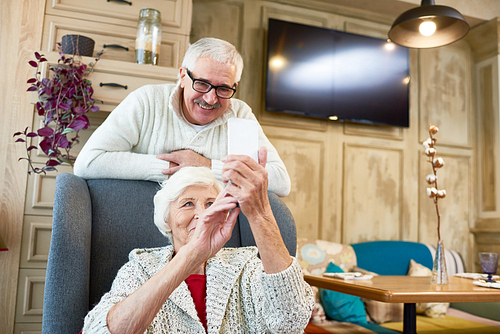 Tech-savvy elderly woman sitting on cozy armchair while taking selfie with her handsome husband with help of smartphone, stylish interior of living room on background
