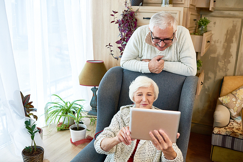 Beautiful aged woman wearing knitted cardigan looking through photos on digital tablet while spending winter day with handsome husband at home, stylish interior of living room on background